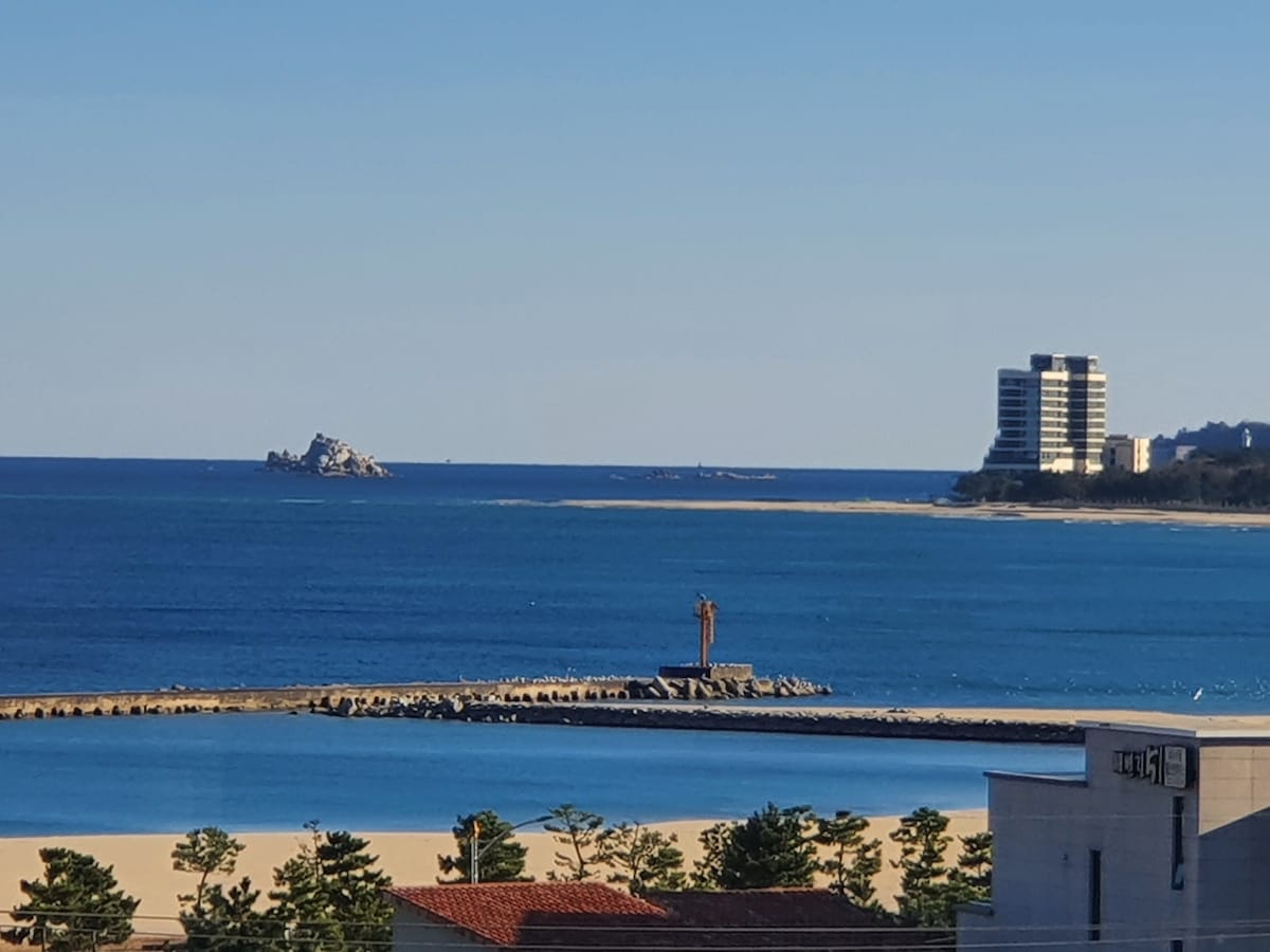 A distant view of the coastline is presented, featuring a calm blue sea meeting a sandy beach. A lighthouse stands on a breakwater, with a few buildings visible nearby. The horizon showcases scattered natural rock formations and residential structures along the shore.