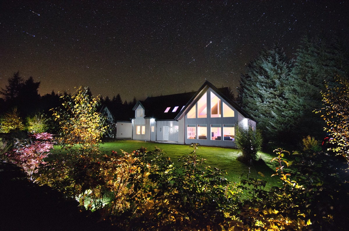 The exterior of a modern eco home is illuminated at night, surrounded by landscaped gardens. Tall trees frame the scene under a starry sky, with bright windows reflecting the natural light inside. Soft landscape lighting highlights the colorful foliage in the foreground.
