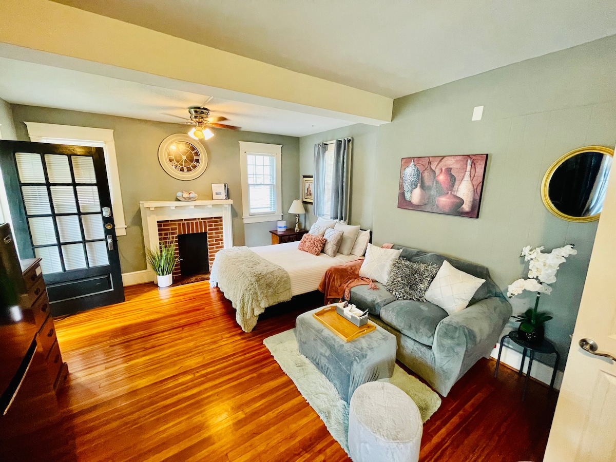 The studio features a spacious bed dressed in soft linens, situated near a cozy seating area with a sectional sofa. A decorative fireplace is present, with art displayed on the wall. Natural light enters through large windows, highlighting the wooden flooring.