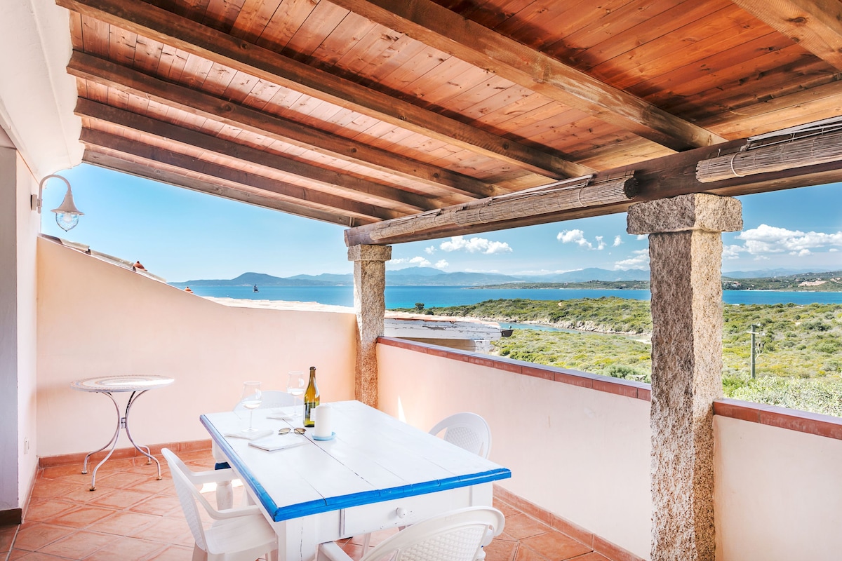 A veranda is presented with a white table and chairs, providing a serene view of the sea and surrounding landscape. The wooden ceiling and stone columns add a rustic touch. Natural light enhances the outdoor seating area, perfect for dining or relaxing.