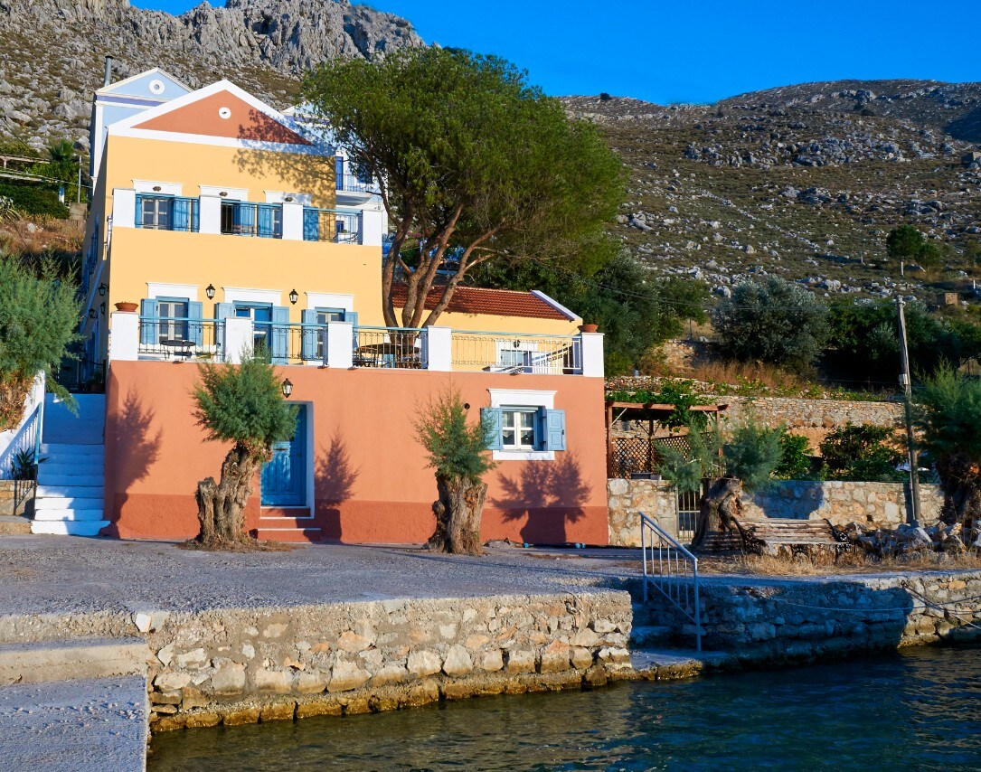 The seafront house is positioned along the water's edge, featuring two levels with colorful facades. Olive trees frame the entrance, while a stone path leads to the waterfront. The surrounding hills and greenery create a serene backdrop, enhancing the peaceful coastal setting.