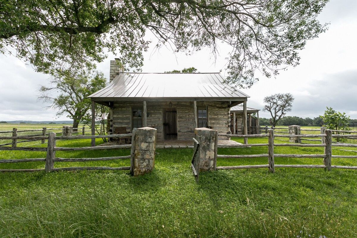 A rustic log cabin is showcased amidst lush green grass and trees, framed by a wooden fence. The structure features a metal roof and a covered porch, contributing to its historic charm. A peaceful country setting is evident with open fields in the background.