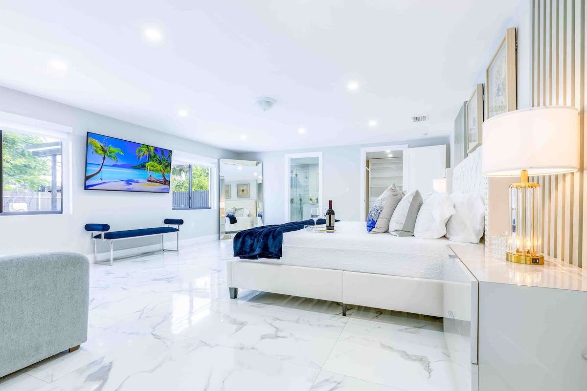 A spacious bedroom features a large bed adorned with decorative pillows. A flat-screen television displays a serene beach scene. Natural light fills the room through multiple windows, and a comfortable seating area with a sofa is present on one side.