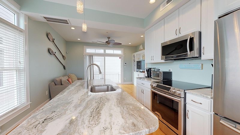 A modern kitchen with a large island features a stainless steel sink and sleek appliances. Light-colored cabinets complement the granite countertop. Bright natural light fills the space from large windows, revealing a cozy seating area in the background adorned with decorative items.
