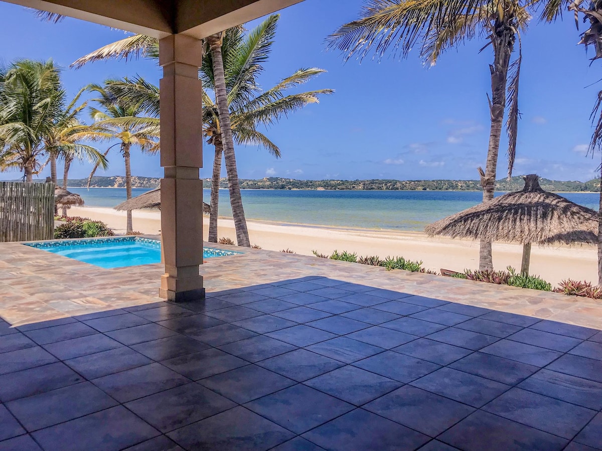 An open patio area features large tiled flooring, providing a view of a tranquil beachfront and lagoon. Lush palm trees frame the scene, while a private swimming pool is situated nearby. Clear blue skies and soft sandy shores enhance the serene coastal environment.