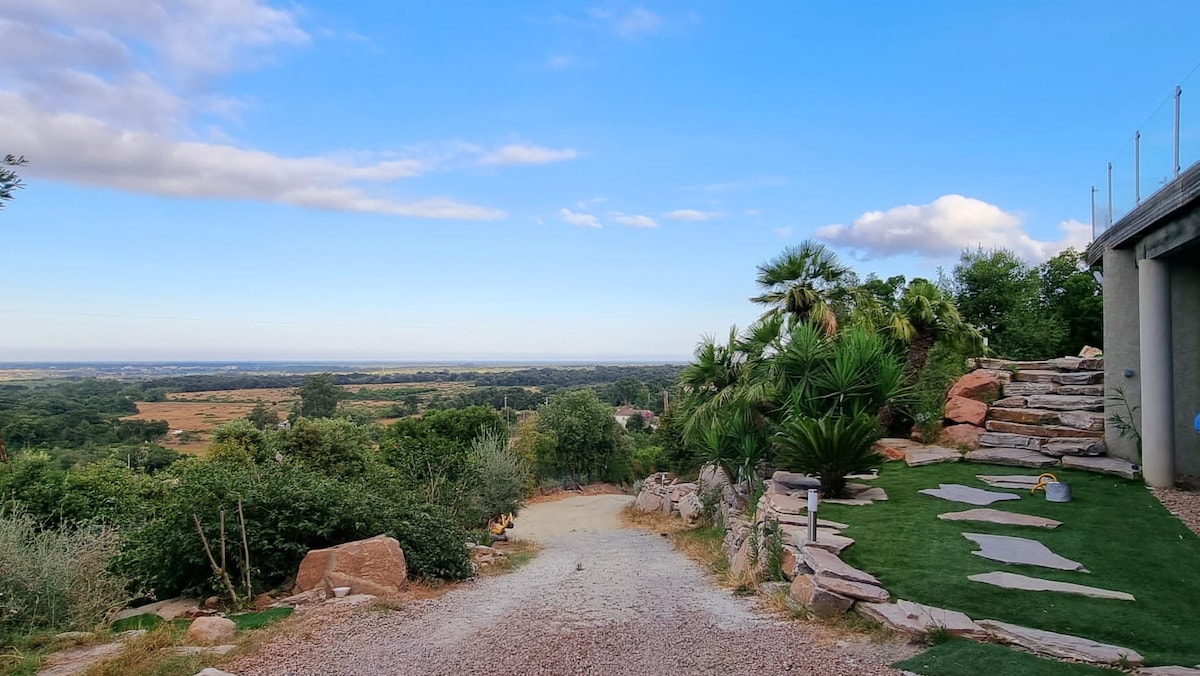 A winding pathway leads through a landscaped area featuring greenery and natural stones. A panoramic view of the eastern plains stretches into the distance under a clear blue sky, with a few scattered clouds enhancing the serene landscape.