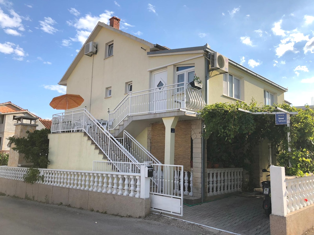 A multi-level family apartment is showcased from the exterior, featuring a spacious terrace adorned with an orange umbrella. A white railing encircles the entrance, where stairs lead to the upper floor, complemented by lush greenery along the façade. Air conditioning units are visible.