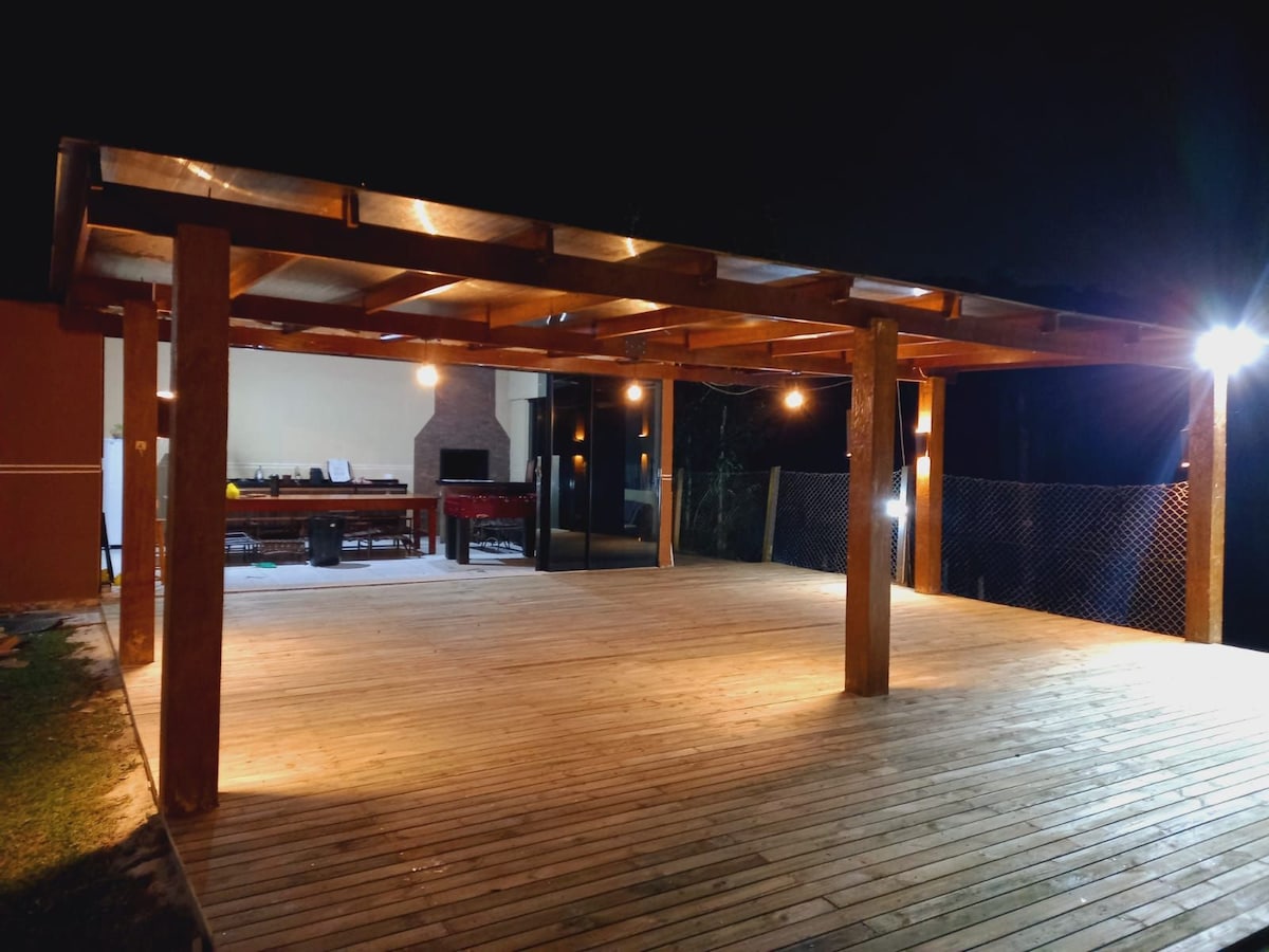 An expansive outdoor deck is illuminated by bright lights, showcasing a wooden structure with a spacious wooden floor. A covered area is visible at the back, hinting at barbecue facilities, while the surrounding area is enclosed by a low fence.