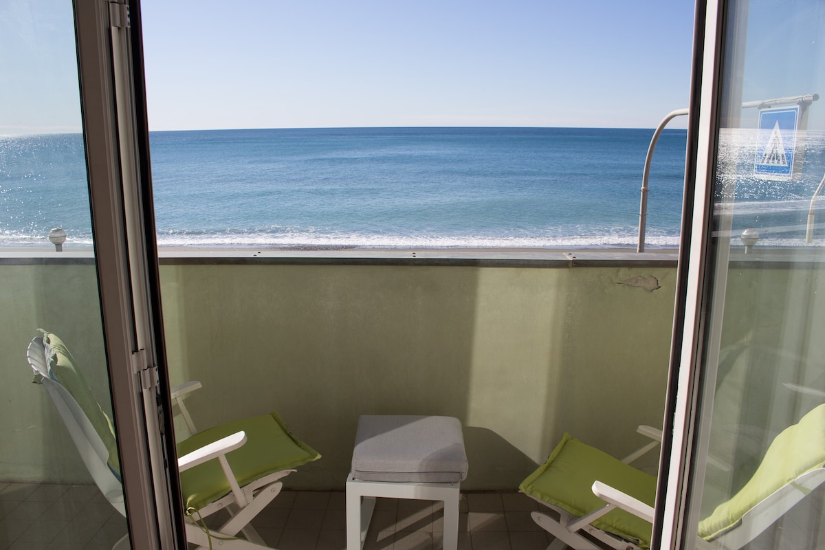 A balcony view captures the serene sea extending to the horizon. Two light green chairs and a small table are positioned on the terrace, inviting relaxation. The clear blue sky meets the ocean, reflecting sunlight on the water’s surface.