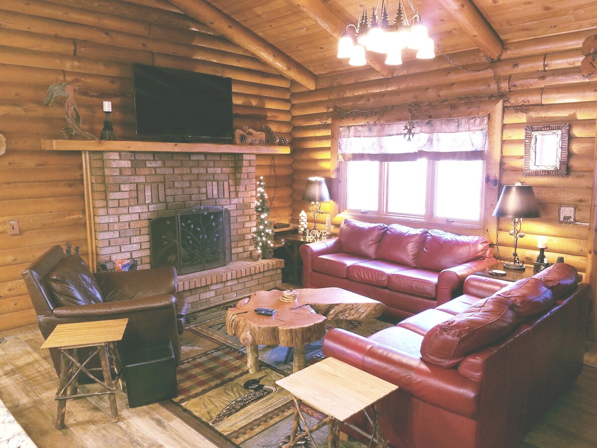 A cozy living area is highlighted by warm wooden logs and features a large stone fireplace. Two red leather sofas are arranged around a central coffee table made of wood. A flat-screen TV is mounted above the fireplace, with soft lighting provided by lamps on either side.