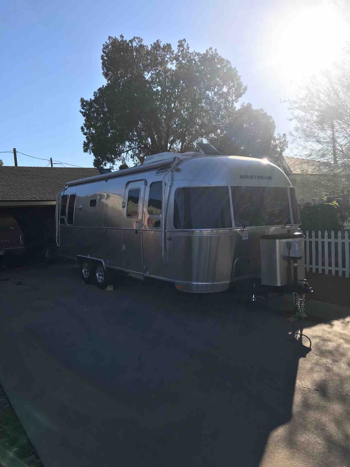 A shiny silver Airstream trailer is parked alongside a house. The bright sun casts a glow over the trailer, highlighting its curved design and polished exterior. Lush greenery is visible in the background, contributing to the outdoor ambiance.