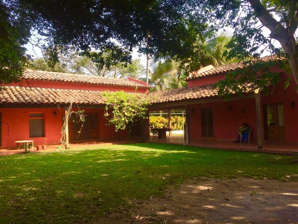 The image showcases a vibrant red house surrounded by a spacious lawn. A patio area is visible, featuring a tile roof and wooden beams. Lush green trees provide shade, and a pathway leads to the entrance. Natural light filters through the foliage, creating an inviting outdoor space.