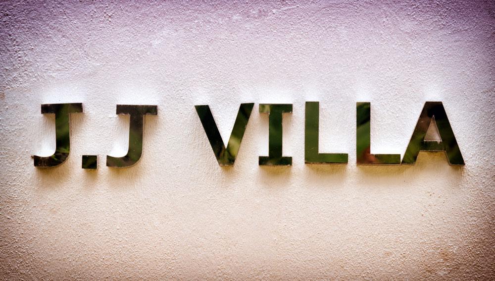 The villa's name is displayed in polished metallic letters against a textured, neutral-toned wall. The design merges seamlessly with the surrounding environment, providing a subtle and elegant introduction to the property.