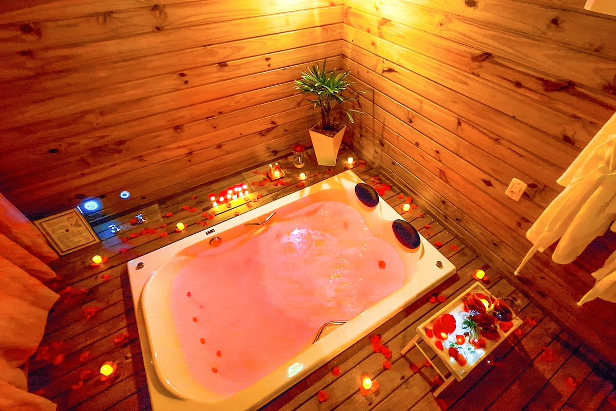 A private hydro massage tub is surrounded by soft lighting and adorned with rose petals. Wooden walls add a warm touch, while a small potted plant enhances the natural feel. The space is designed for relaxation and tranquility.