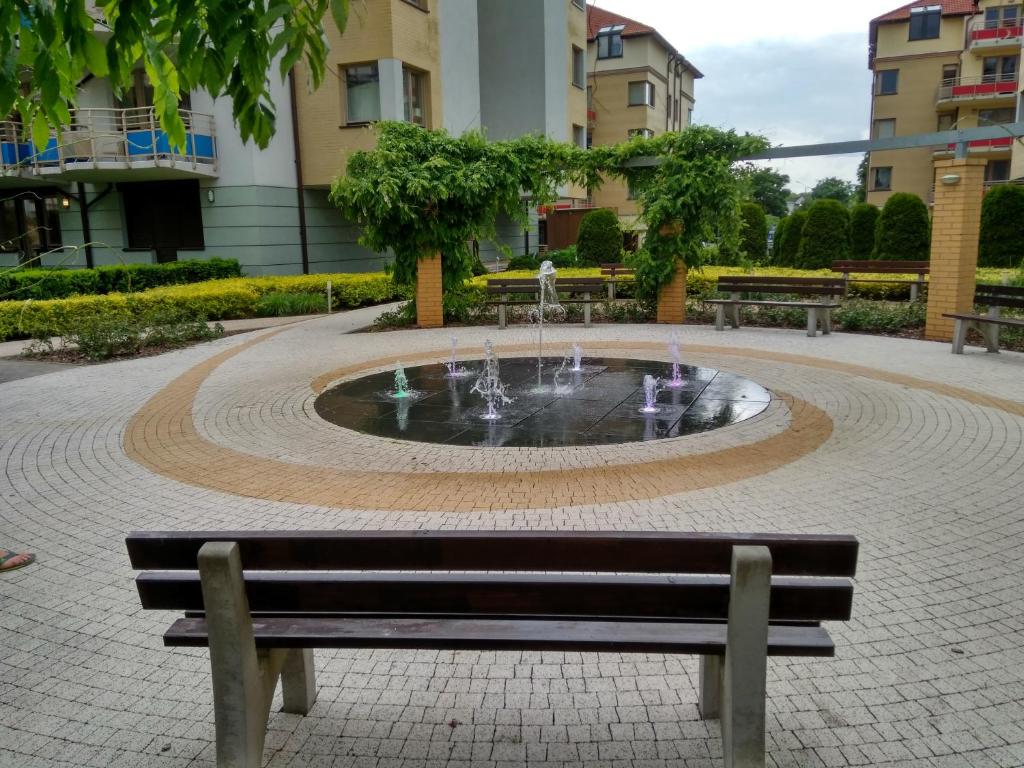A peaceful courtyard features a geometric fountain at its center, surrounded by a stone path and vibrant greenery. Benches provide seating around the fountain, with a backdrop of residential buildings enhancing the tranquil atmosphere.