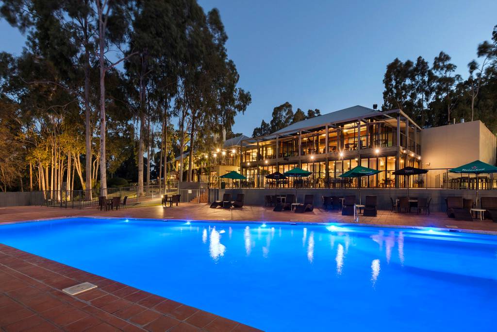 The outdoor pool area is illuminated in a calming blue light, surrounded by lounge chairs. The resort building, featuring large windows and a spacious balcony, is visible in the background, framed by tall trees. Warm lighting from the building enhances the tranquil setting.