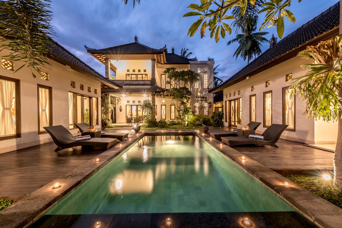 The exterior of the villa is shown, featuring a tranquil swimming pool illuminated by soft lighting. Surrounding the pool are sun loungers, and the lush greenery adds to the relaxing atmosphere. The traditional Balinese architecture of the villa is visible, creating a harmonious blend with nature.
