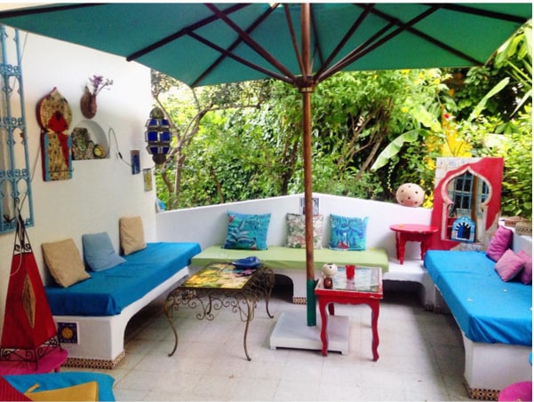 A shaded outdoor seating area is provided, featuring blue and green lounging cushions placed on curved benches. A colorful table sits in the center, surrounded by vibrant decorations on the walls and lush greenery in the background.
