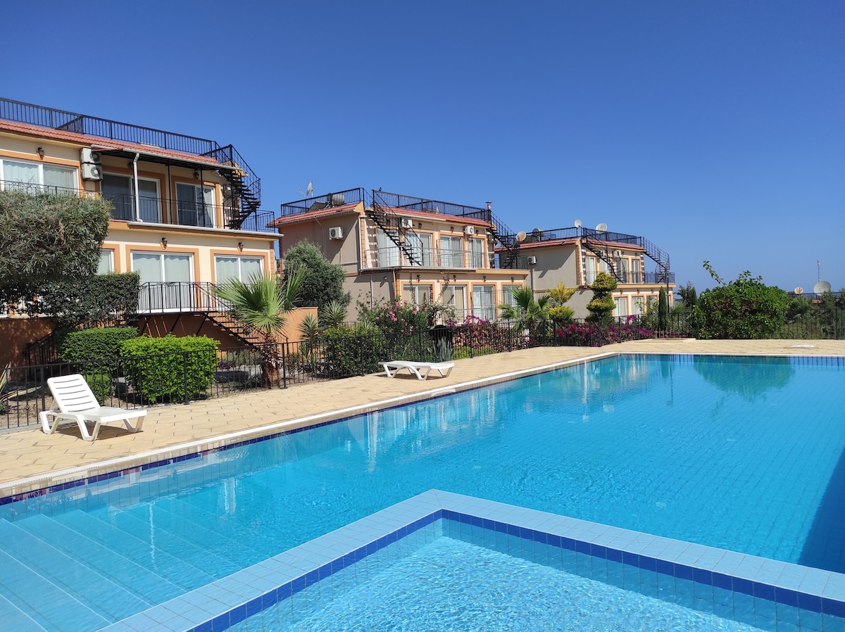 A sparkling blue pool reflects the vibrant sky, surrounded by landscaped gardens and colorful flowers. Multiple apartment units with balconies and staircases are visible in the background, providing a welcoming setting for relaxation and leisure.