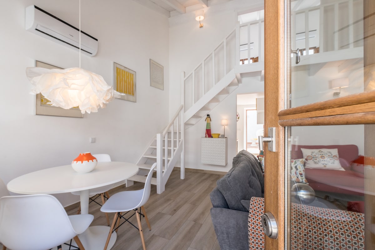 A bright open-plan living area features a round dining table with white chairs and a decorative centerpiece. A cozy couch in a muted tone is adorned with colorful cushions. Soft lighting enhances the white walls, while an air conditioning unit is mounted above.