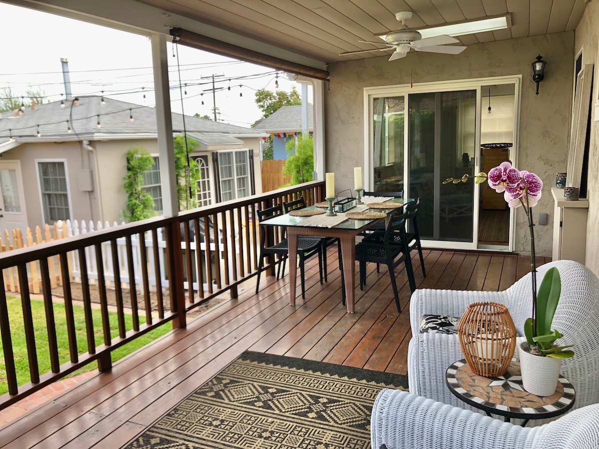 The spacious outdoor deck features a wooden floor with a dining table set for four. A comfortable seating area includes a white sofa and a decorative coffee table with a plant and woven basket. Lush greenery is visible in the background.
