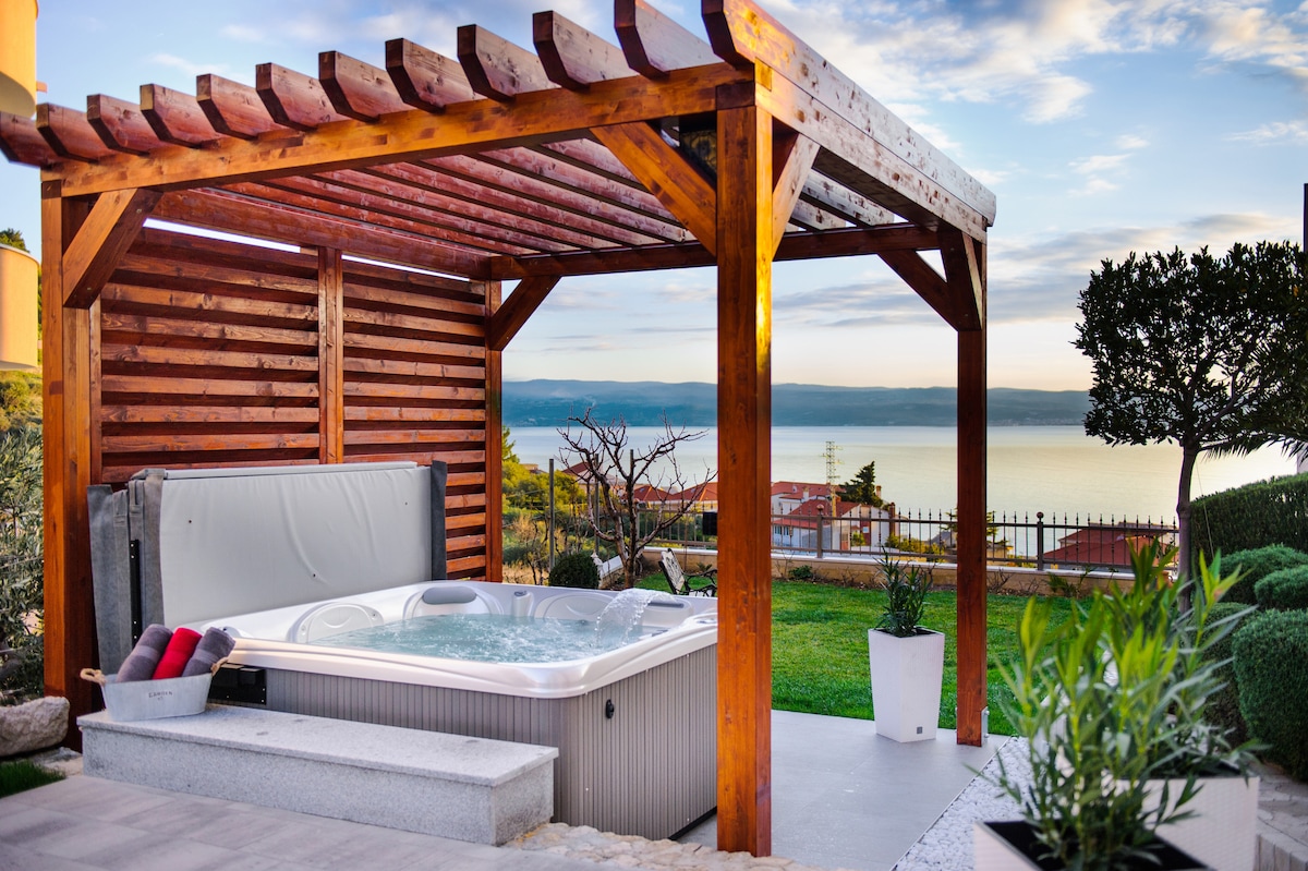 An outdoor hot tub is positioned under a wooden pergola, with a view of the sea in the background. Surrounding greenery adds a natural touch, complemented by neatly trimmed hedges and potted plants. The sky is clear, creating a serene atmosphere.