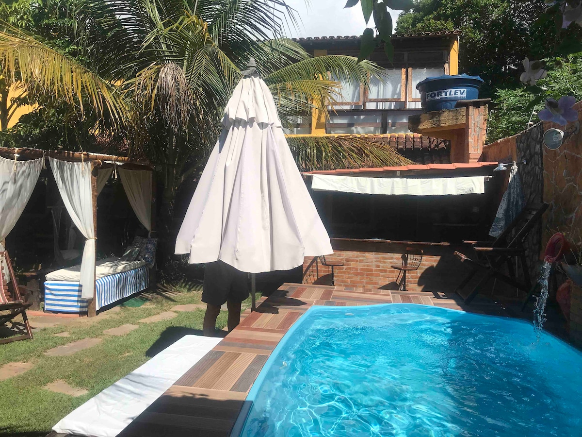 A spacious outdoor area features a refreshing pool surrounded by lush greenery. A gazebo stands nearby, offering shade, while lounge chairs and a decorative parasol enhance the relaxing atmosphere. The vibrant colors of the landscaping complement the inviting blue of the water.