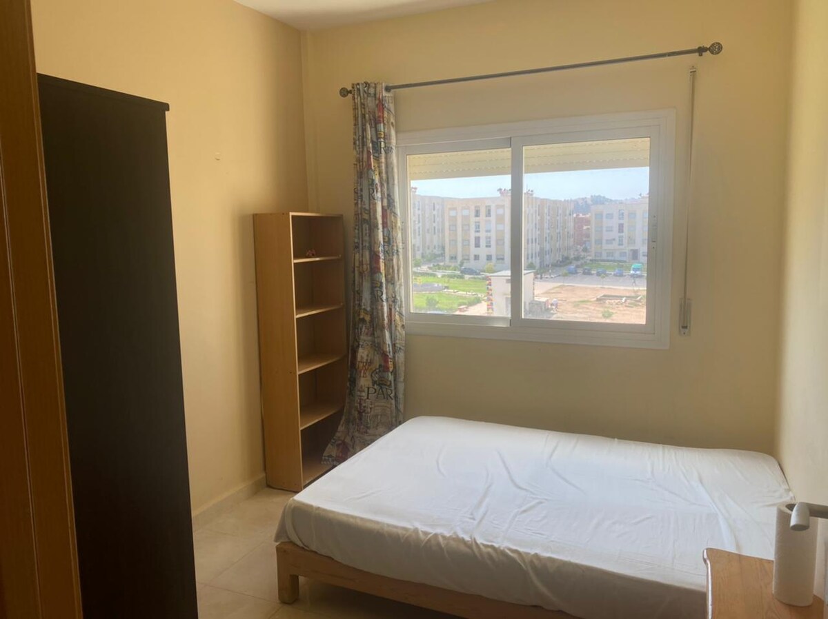 A bright bedroom features a spacious bed with a minimalist design. A large window with sheer curtains allows natural light to fill the space, revealing a view of the surroundings. A wooden wardrobe and a shelf provide additional storage options.