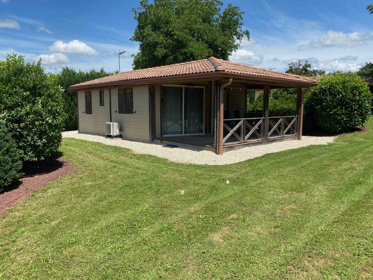 A single-story chalet is nestled within green landscaping, featuring a covered terrace with wooden railings. Large windows provide natural light, while gravel pathways lead around the exterior. The sloped roof and colorful greenery create a serene setting.