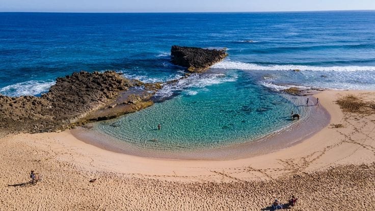 A serene beach scene captures gentle waves lapping against a sandy shore. Clear, calm waters form a circular cove, bordered by rocky outcrops. People can be seen walking along the water's edge, while footprints mark the sandy expanse.