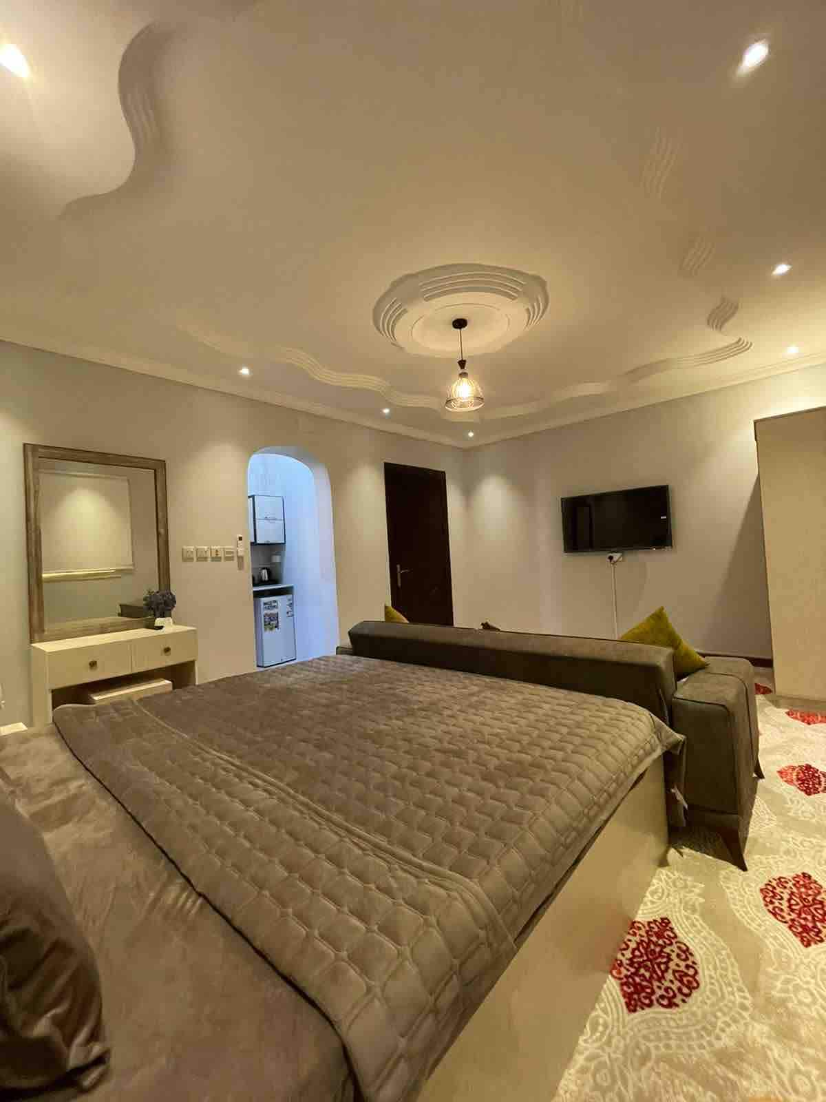 The room features a spacious bed with a quilted cover, complemented by a comfortable sofa and a sleek dresser. A wall-mounted television is visible, along with soft lighting from ceiling fixtures that enhance the room's modern design.