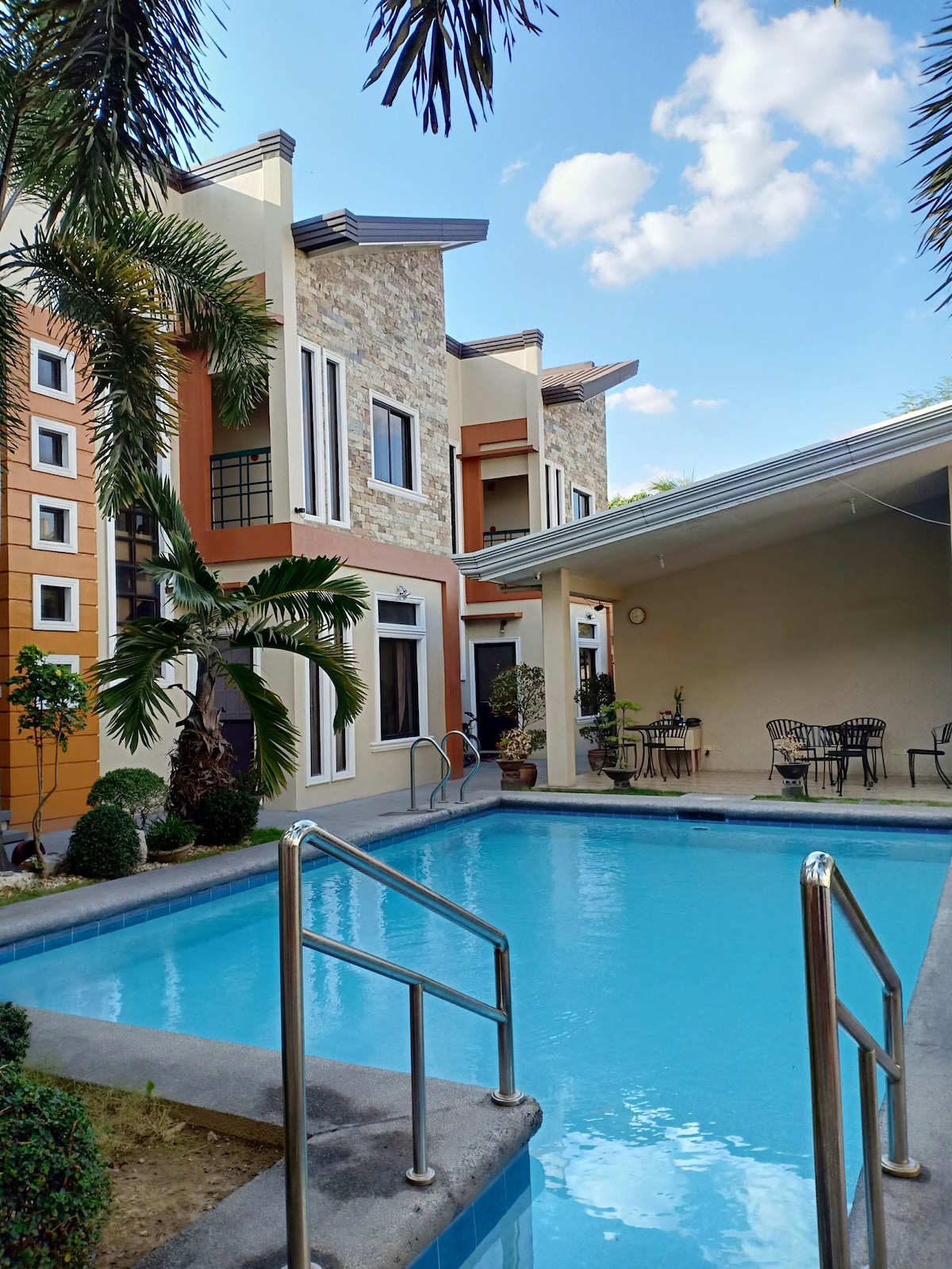 A well-maintained pool is framed by lush greenery, with elegant palm trees nearby. The surrounding residential building features a mix of stone and colorful exterior walls. A shaded seating area with tables and chairs is also visible alongside the pool.