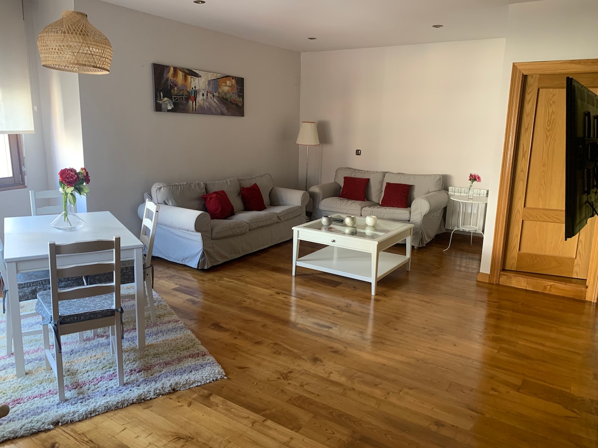 A spacious living area features a light wooden floor and large windows allowing ample natural light. Two sofas, adorned with red cushions, face a white coffee table. A dining set with four chairs is positioned next to a colorful rug, providing a comfortable and inviting environment.