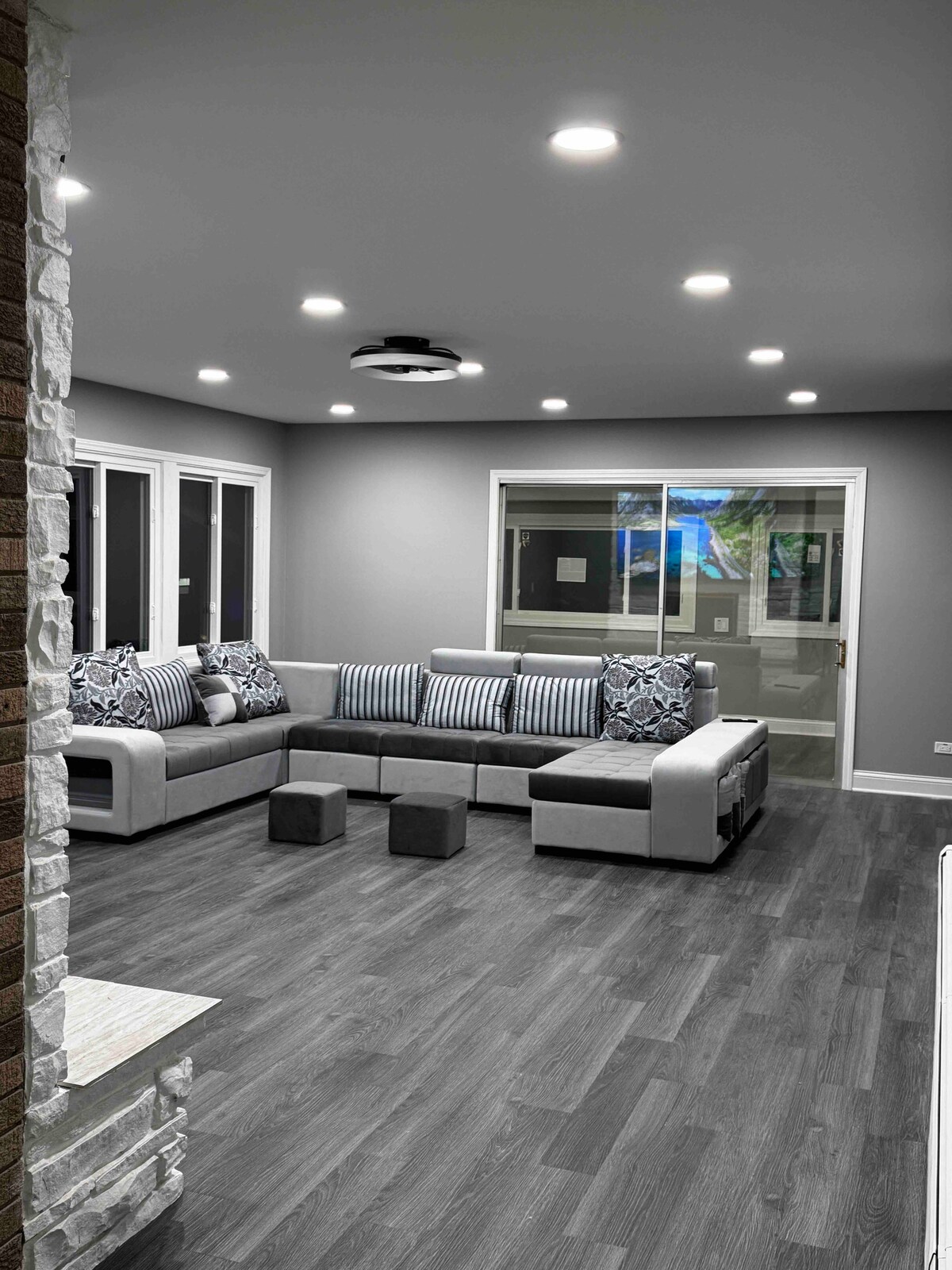 A spacious living area features a modern sectional sofa adorned with patterned cushions. Natural light is welcomed through large windows, illuminating the sleek wood flooring. Recessed lighting enhances the ambiance, while a projector screen is subtly integrated into the ceiling.