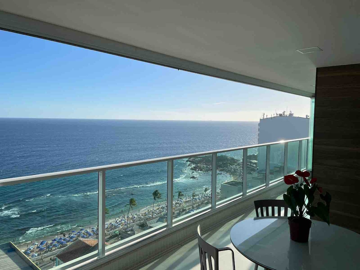 A panoramic view of the ocean is visible through large glass sliding doors, showcasing bright blue waters and a sandy beach below. A round dining table with a potted plant is positioned on the balcony, providing a serene space to enjoy the scenery.
