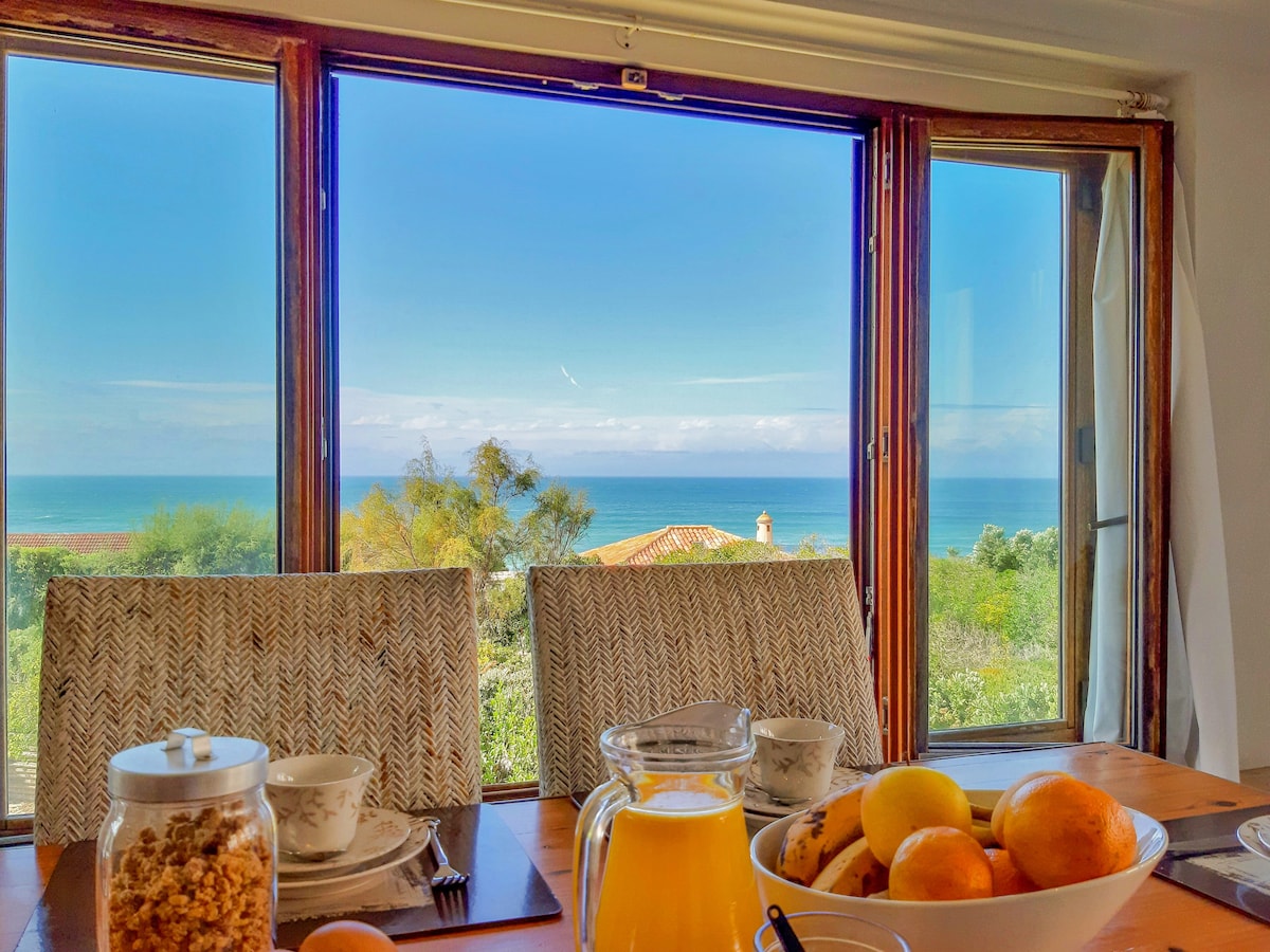 A dining table is set with bowls and glassware, positioned in front of a large window offering panoramic views of the ocean and sky. A colorful fruit bowl sits at the center, inviting natural light into the airy space.