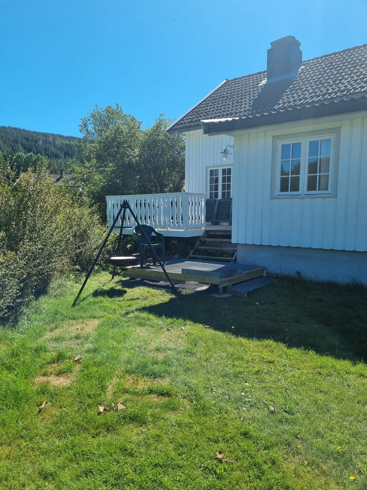 A sunny outdoor area features a white house with a gabled roof, surrounded by greenery. A wooden swing set is positioned on a grassy lawn, with a small deck accessible from the house, which includes a few outdoor chairs for seating.