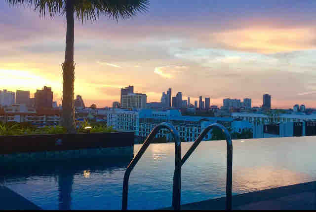 An infinity pool is visible on the rooftop, reflecting the colorful hues of a sunset. City skyline silhouettes appear in the background, framed by a palm tree. The tranquil ambiance suggests a serene retreat.