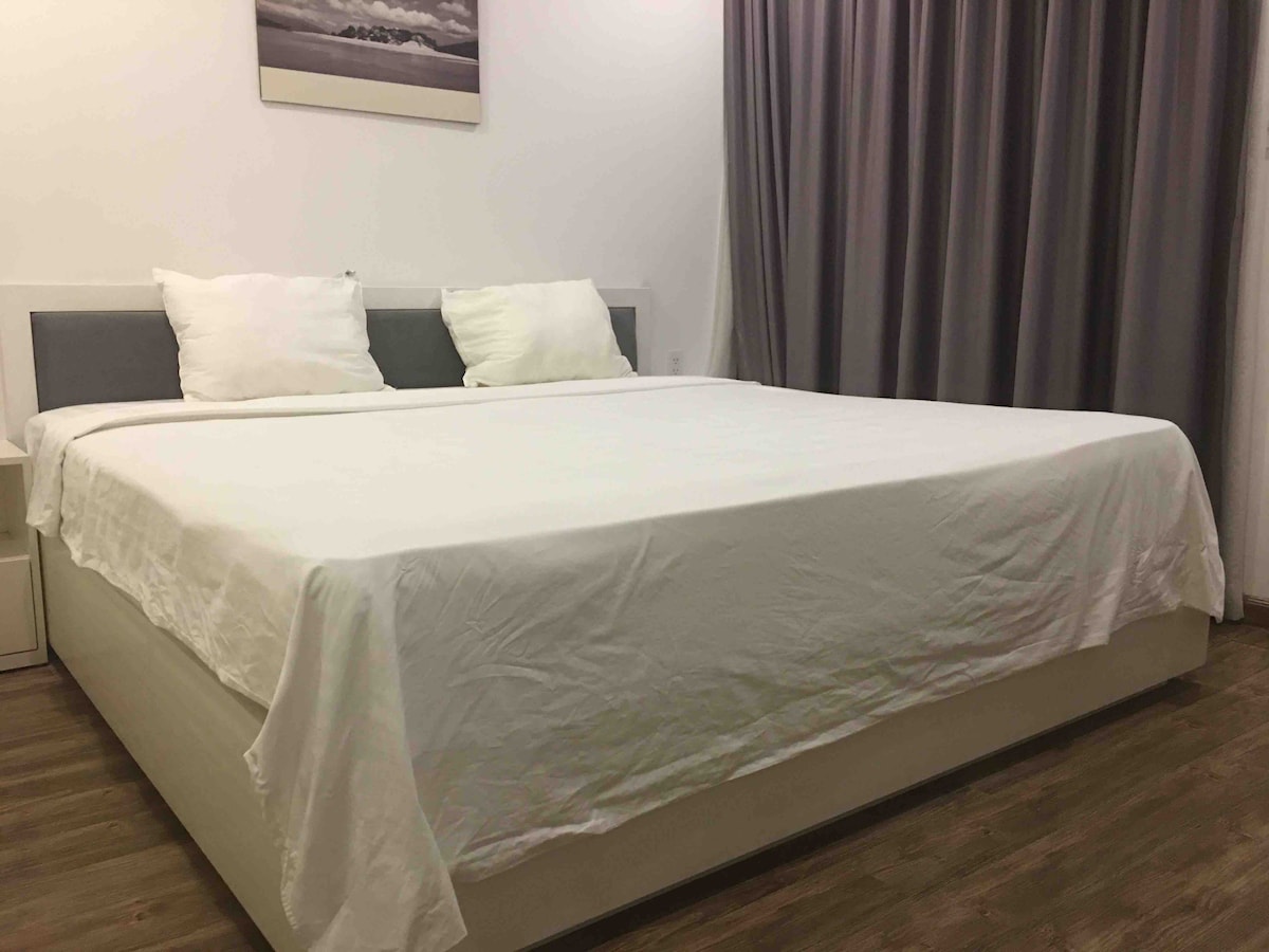 A spacious bed with crisp white bedding is positioned against a simple headboard. Two decorative pillows rest at the back. Light-colored curtains frame a window, allowing natural light to softly fill the room, creating an open and airy feel.