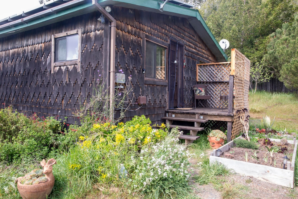 A rustic wooden exterior is complemented by a welcoming entryway featuring steps leading up to a covered porch. A vibrant flower garden filled with seasonal blooms sits in front, with visible patches of vegetables in garden beds nearby, reflecting the farm's sustainable practices.