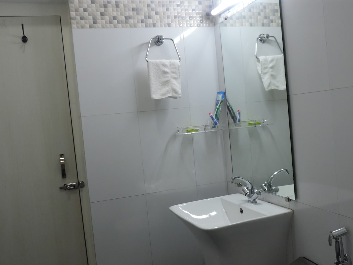 A modern bathroom features sleek white tiles and a compact sink with a contemporary faucet. Above the sink, a large mirror reflects the space, while a shelf holds toiletries. Two white towels hang neatly on a silver rack, enhancing practicality.