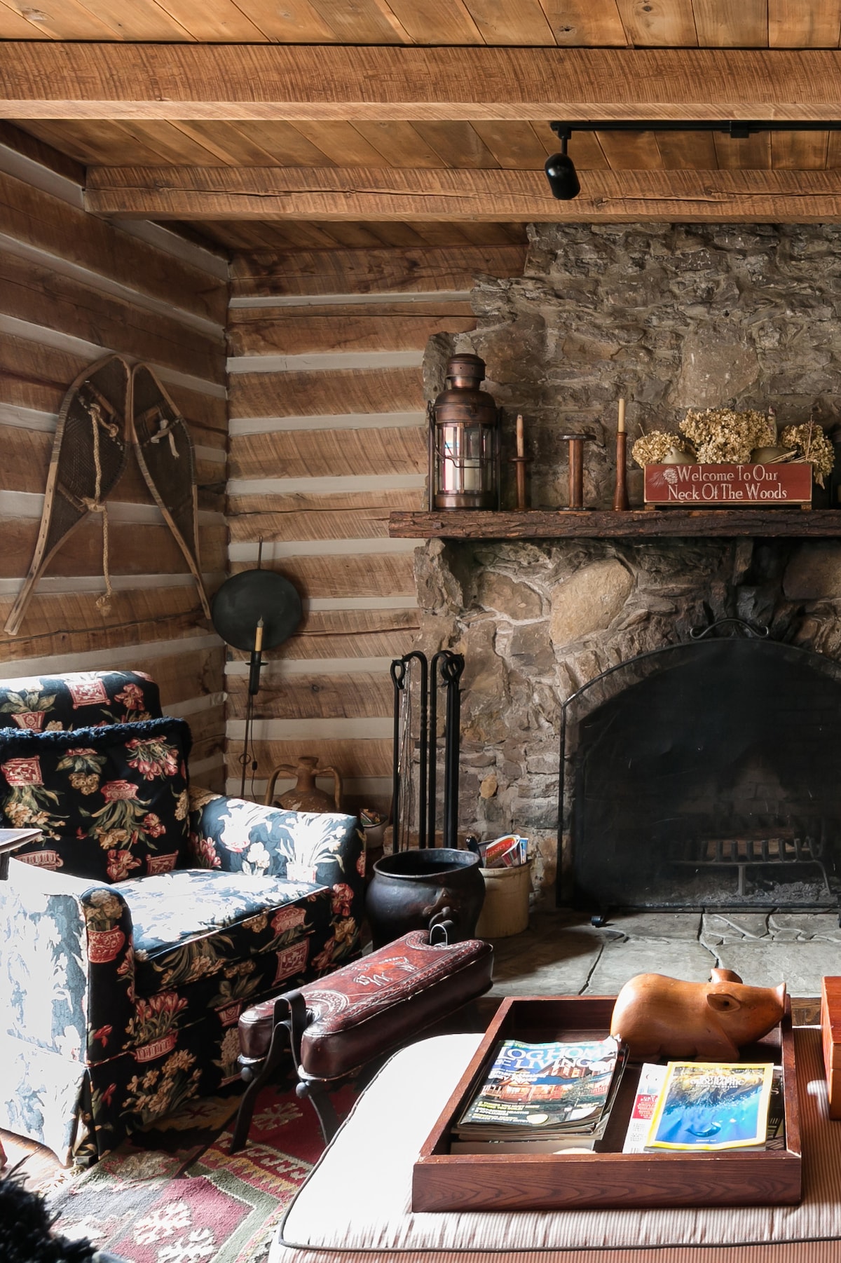 A cozy living space features rustic wooden beams above and a traditional stone fireplace as its focal point. Floral-patterned armchairs provide comfortable seating, while a wooden coffee table displays a selection of magazines. Antiques and decorative items add character to the room.