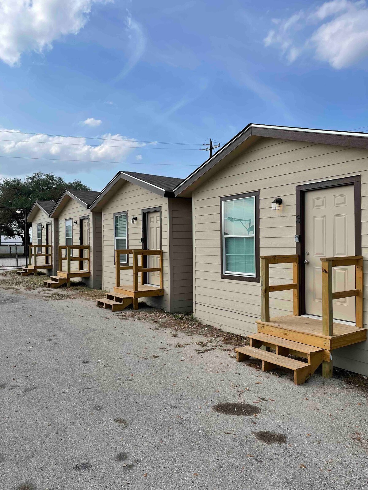 A row of identical cabins features light-colored exteriors with dark trim. Each cabin is equipped with a small wooden porch and a single door with a light fixture above. Green windows allow natural light into the spaces, and the sky above is partly cloudy.