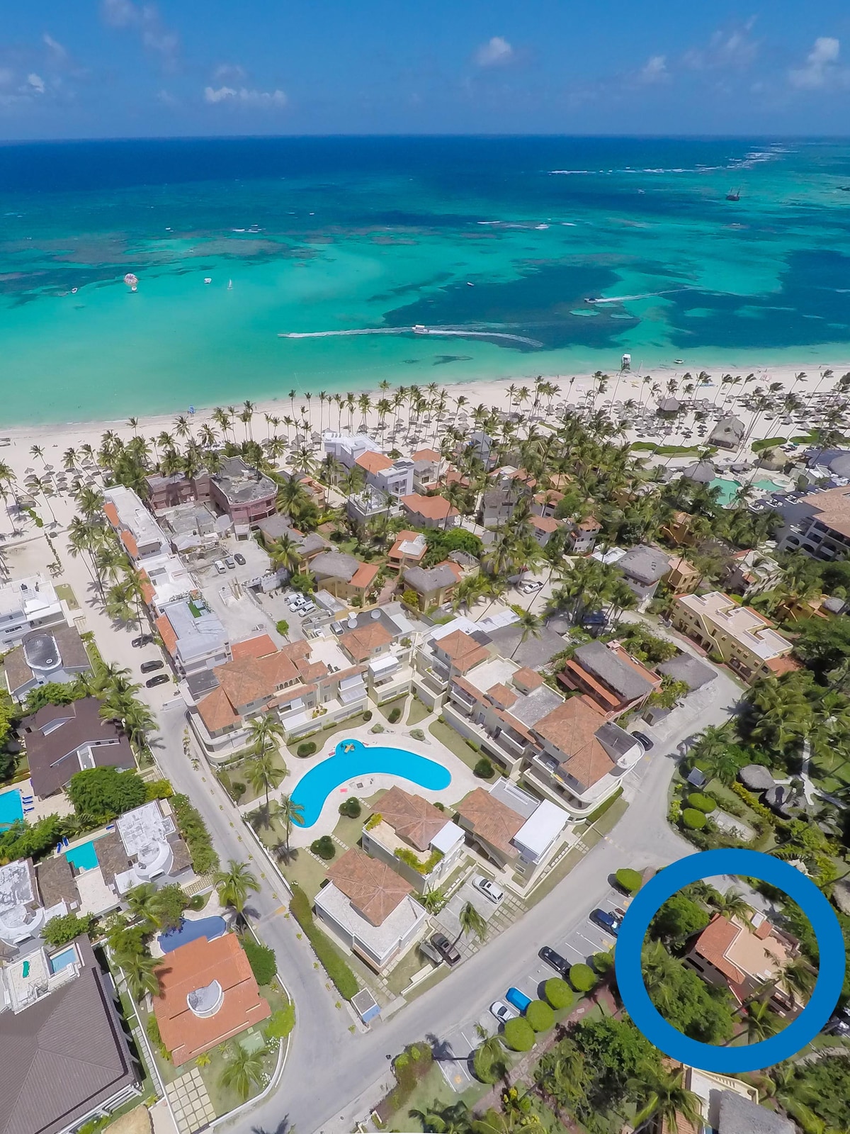 An aerial view captures a coastal neighborhood alongside a vibrant beach. Lush greenery and palm trees surround the Mediterranean-style houses. A clear swimming pool is visible, with the beach's turquoise waters beyond, creating a serene environment near the ocean.