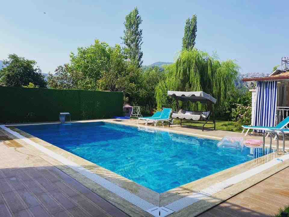 A large swimming pool with crystal-clear blue water is surrounded by a spacious deck. Lounge chairs are positioned along the edges, and a shaded seating area is visible beneath a canopy. Lush greenery can be seen in the background, creating a serene environment.