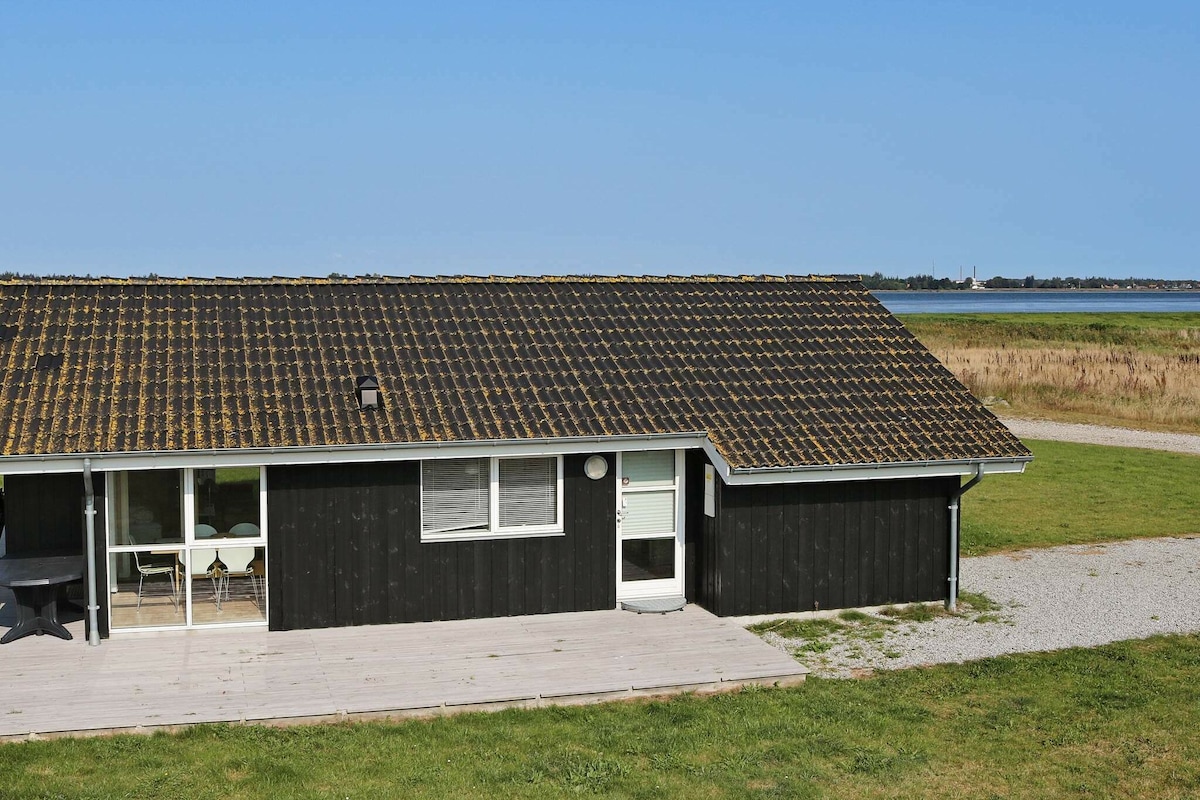 A holiday home is viewed from above, featuring a sloped roof covered with tiles. The exterior is dark wood with a light entrance door. A wooden terrace extends from the house, surrounded by a grassy area and offering a view of the nearby Limfjord.
