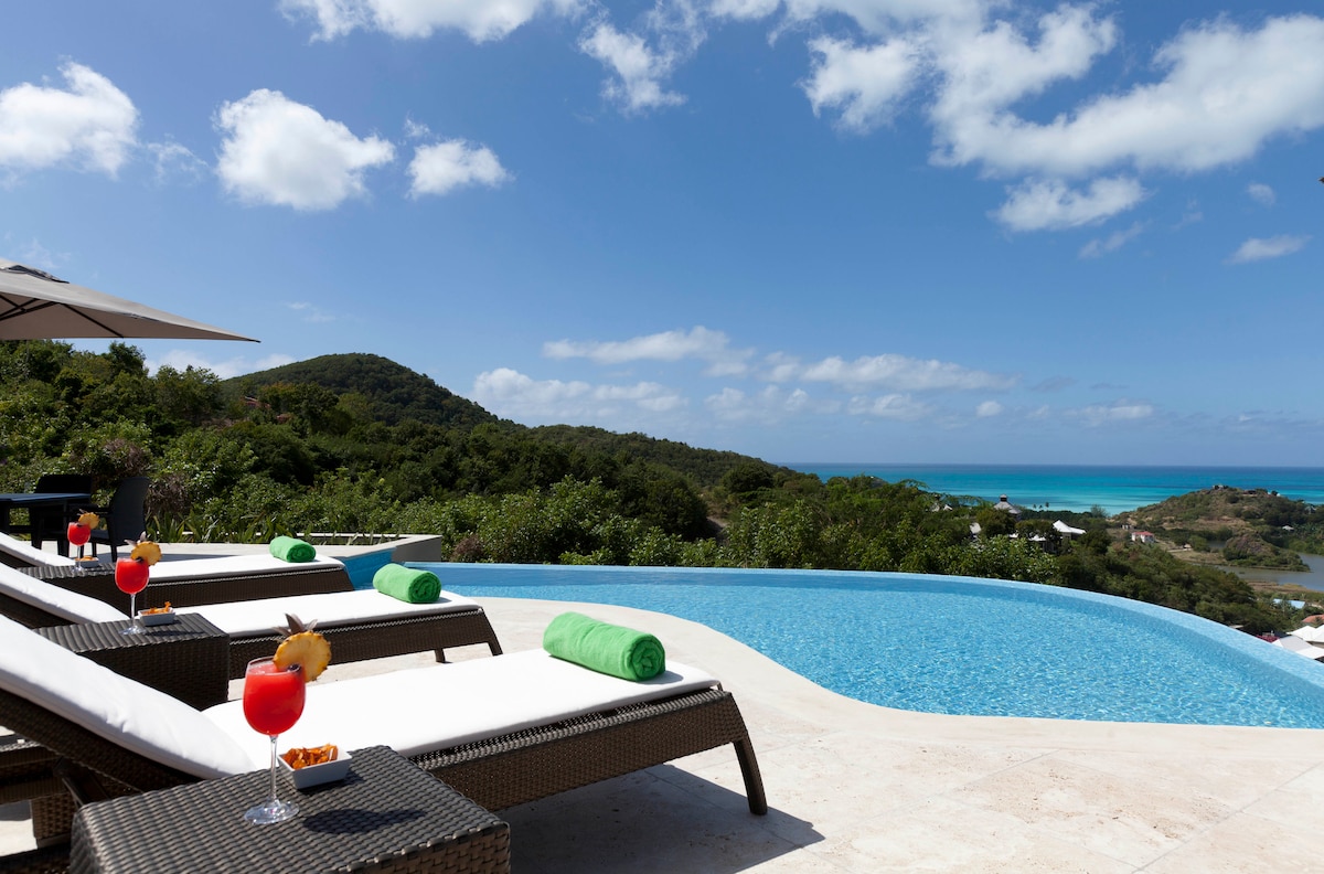 A luxurious infinity pool is framed by lounge chairs with green towels. Tropical drinks are placed on nearby tables, showcasing a stunning view of the Caribbean Sea and lush surroundings under a bright sky with scattered clouds.