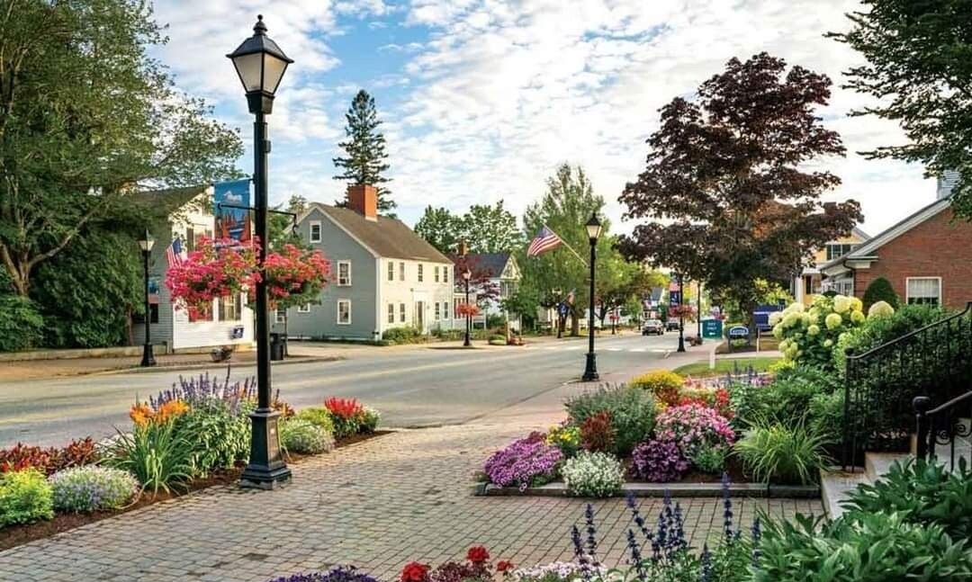 A charming streetscape features flowering gardens and lush greenery lining the walkway, with a cobblestone path leading towards a tree-lined neighborhood. Colorful storefronts and a lamppost accentuate the inviting atmosphere, creating a vibrant scene in the heart of Kennebunk.