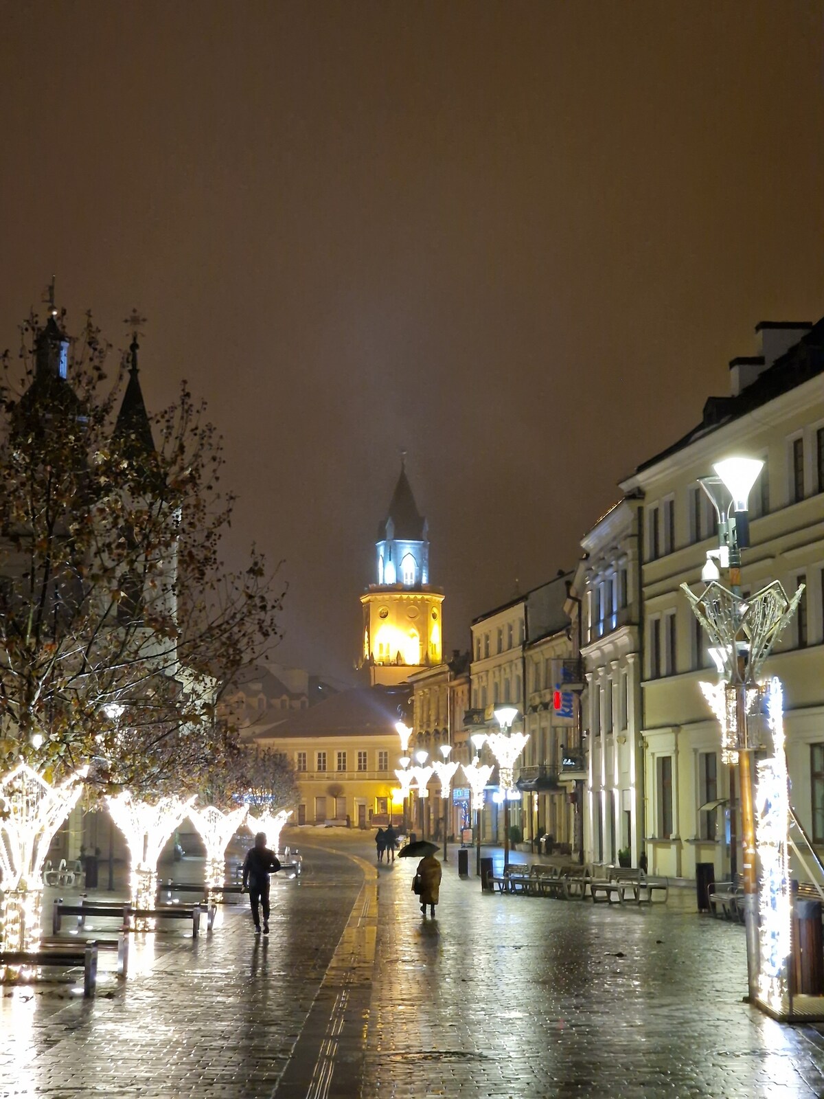 A quiet street in Lublin is illuminated by decorative lights strung around trees and along buildings. A clock tower is visible in the background, indicating the time. The wet pavement reflects the warm light, creating a serene atmosphere on the evening stroll.