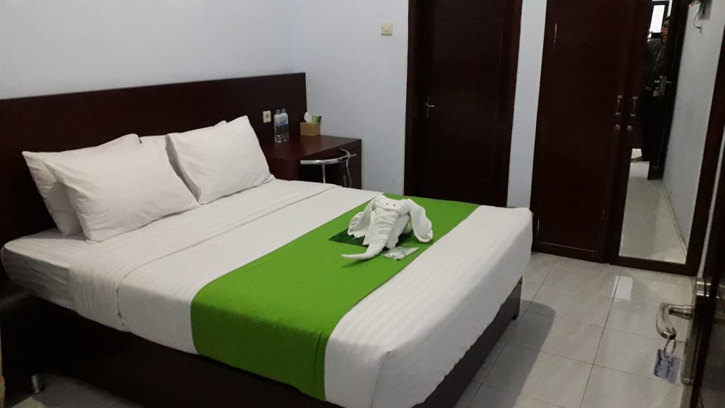 A spacious bed with white linens and a green accent blanket is featured in the room. An elephant towel art is positioned at the foot of the bed. Wooden furniture includes a bedside table with bottles of water. A mirror and door to an en-suite bathroom are visible.